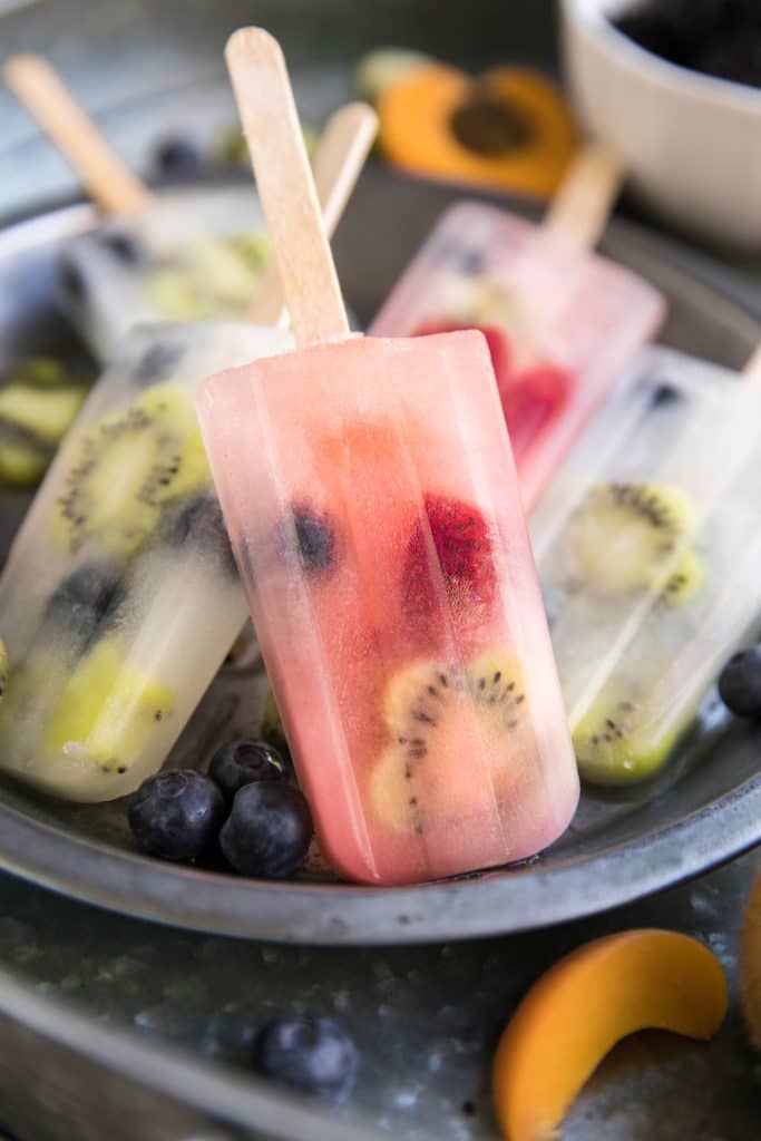 five lemonade and fruit popsicles in a bowl with peach slices and blueberries on the side 