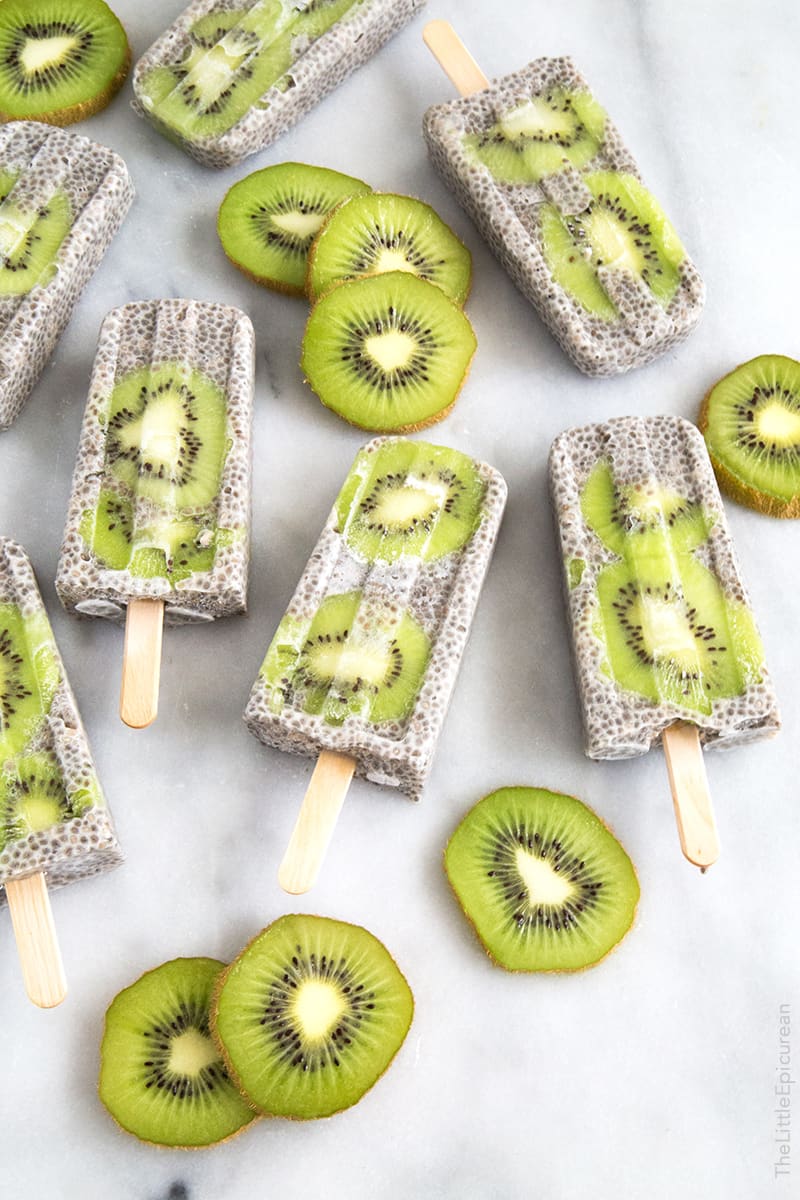 seven chia kiwi popsicles on a marble countertop with kiwi slices on the side 