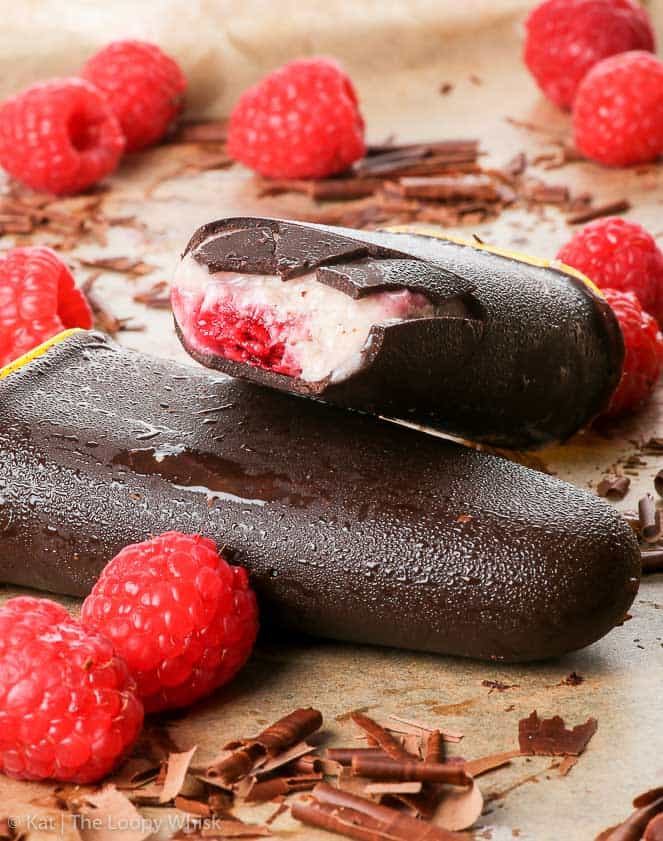two chocolate-covered raspberry popsicles on a parchment paper with fresh strawberries on the side