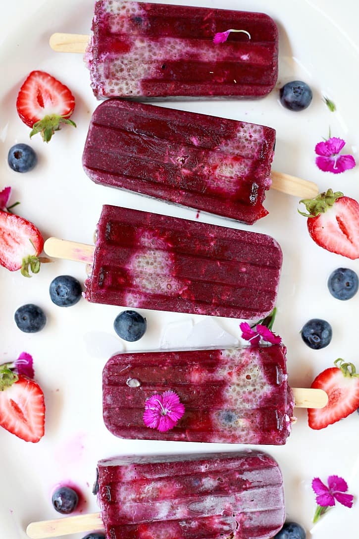 five açaí popsicles on a white plate with strawberries, blueberries, and flowers on the side 