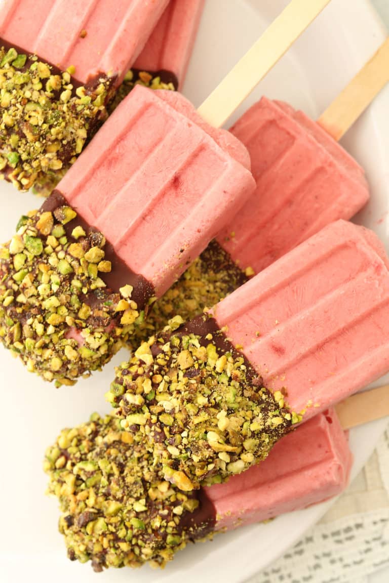six strawberry coconut popsicles on a white plate dipped in dark chocolate and rolled in pistachios 