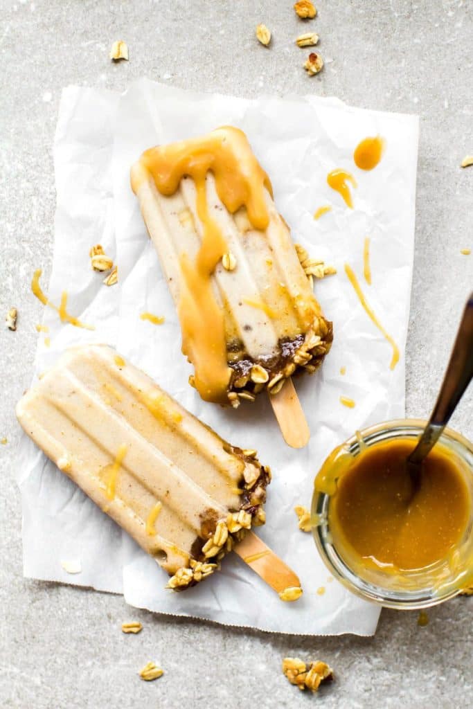 two vegan banoffee ice cream popsicles on a piece of parchment paper with a bowl of homemade toffee sauce with a spoon on the side 