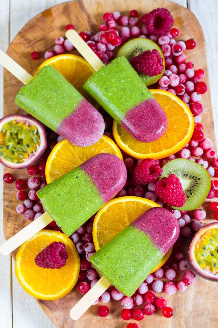 four green and pink smoothie popsicles on a wooden board covered with frozen currants, kiwi, and orange slices 