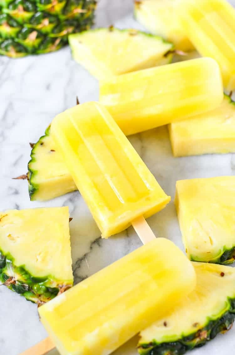 four pineapple lemonade popsicles on a marble countertop with pineapple slices on the side 