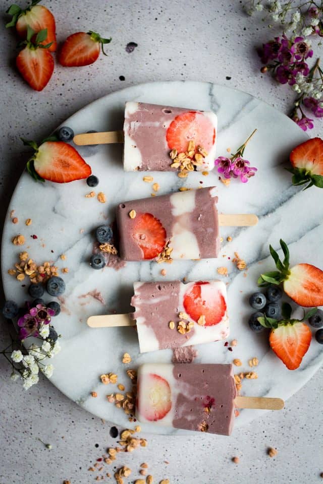 four vegan açaí popsicles on a round marble board with strawberry halves and blueberries on the side 