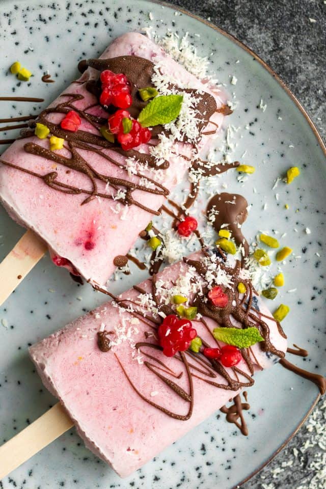two raspberry coconut popsicles on a grey plate covered with chocolate, pistachios, mint leaves, and raspberries 