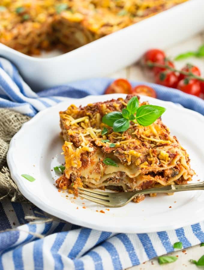 a piece of vegan lasagna on a white plate with a fork on the side and a casserole dish with more lasagna in the background 