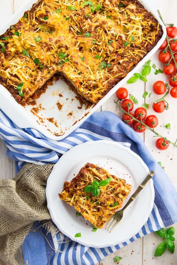 a piece of vegan lasagna on a white plate with a fork on the side and a baking dish with more lasagna in the background 