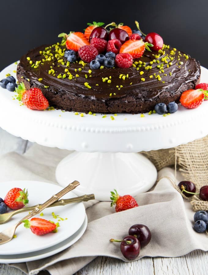 a vegan chocolate cake topped with fresh berries and pistachios on a white cake platter 
