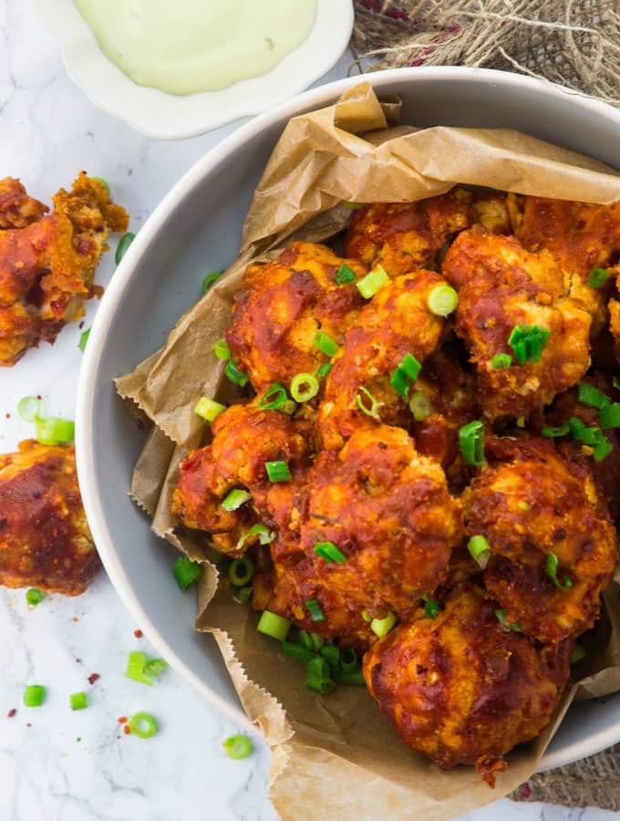 a bowl of cauliflower hot wings on a marble counter top with chopped green onions on top 