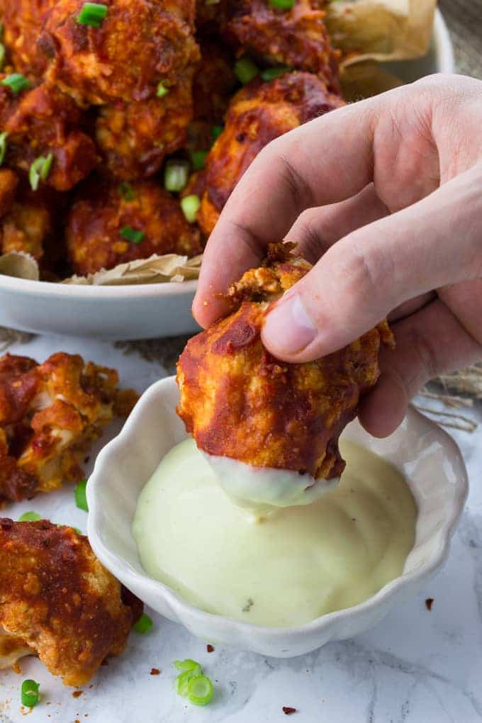 A hand dipping a cauliflower hot wing into a bowl of vegan aioli with more cauliflower wings in the background