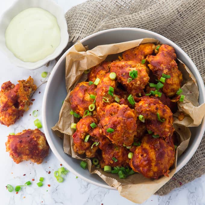 a bowl of vegan cauliflower hot wings on a marble counter top with green onions on top 