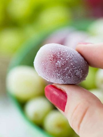 A hand holding a frozen grape with a bowl of more frozen grapes in the background