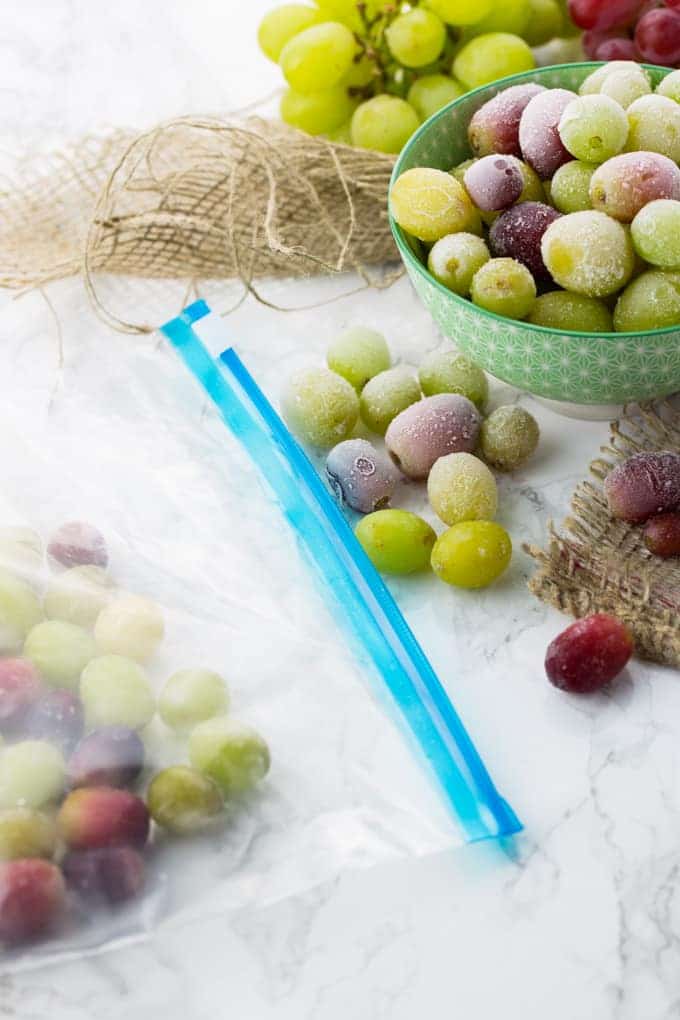 a bowl of frozen grapes on a marble counter top with a ziploc bag of frozen grapes on the side 