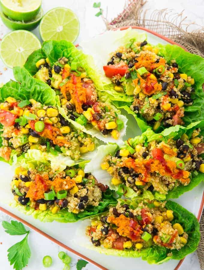 six vegetarian lettuce wraps with quinoa and black beans on a white plate with limes and roughly chopped parsley on the side 