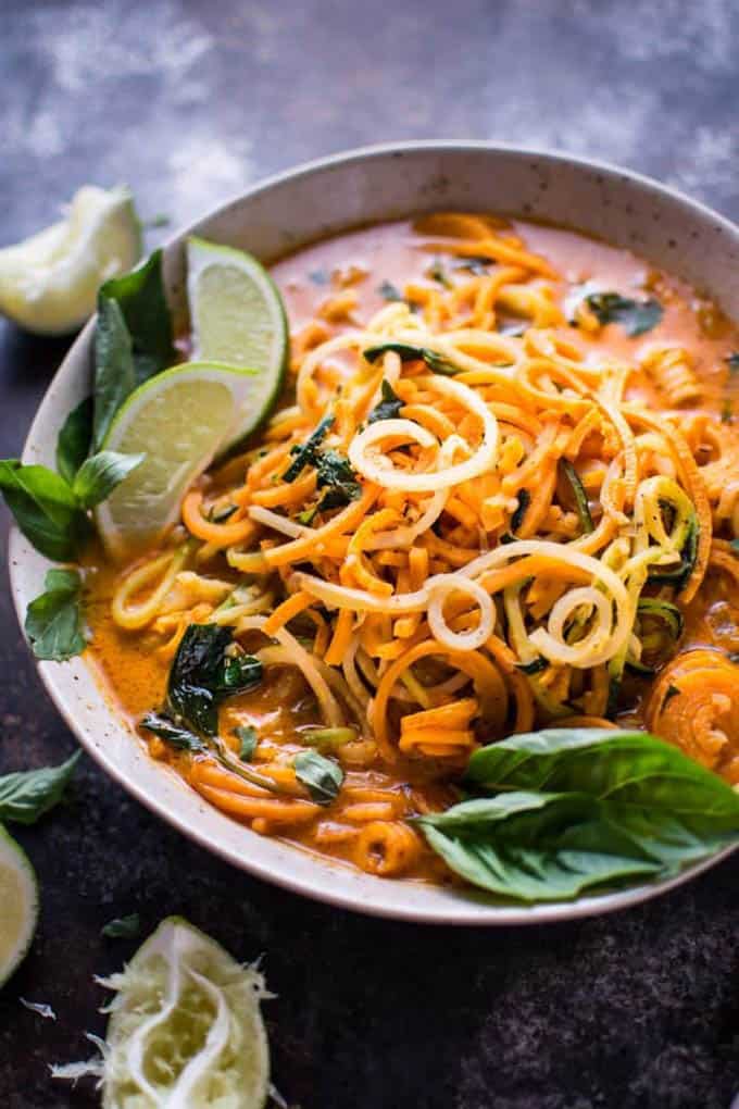 A bowl of Thai lemongrass soup on a grey counter top with lime wedges and basil on top