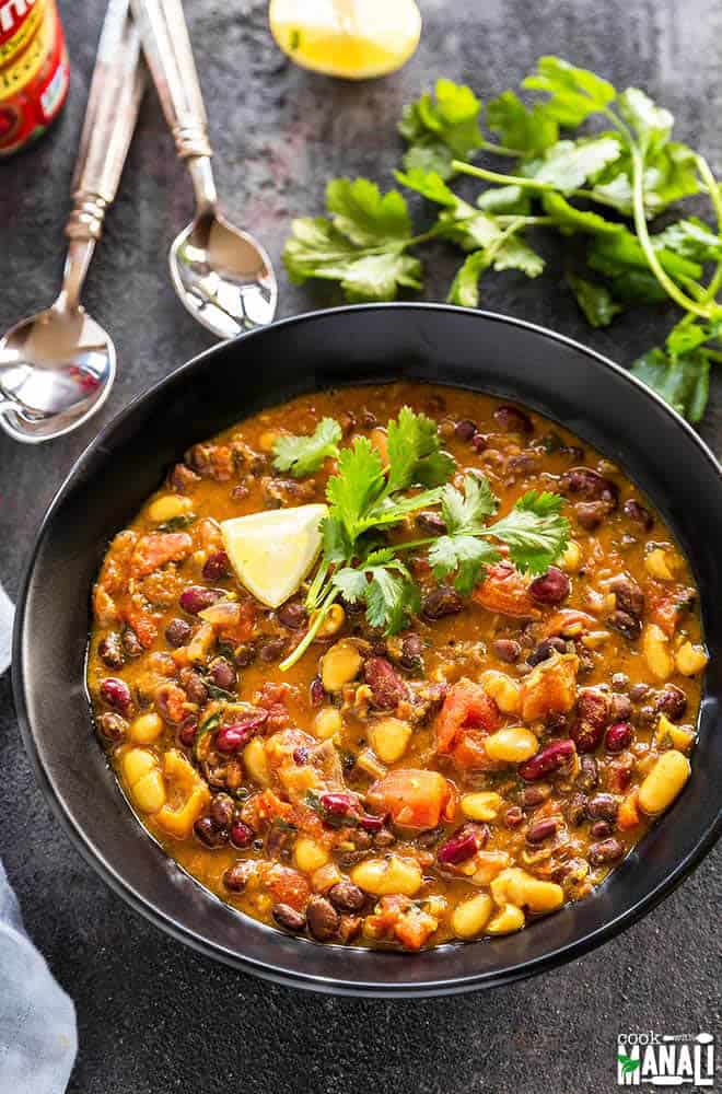 Three bean curry in a black bowl with two spoons and fresh cilantro on the side