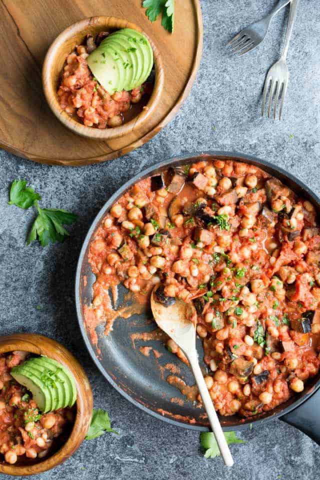 A pan with bean and aubergine casserole with a wooden spoon and two wooden bowls on the side