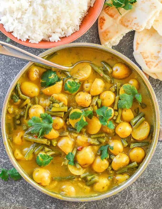 A bowl of green bean potato curry with a bowl of rice and naan bread on the side