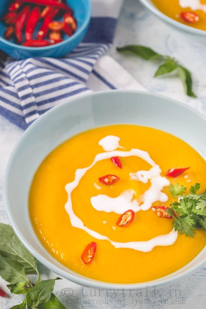 a bowl of vegan pumpkin soup on a white counter top with a blue napkin and a bowl of chilis in the background