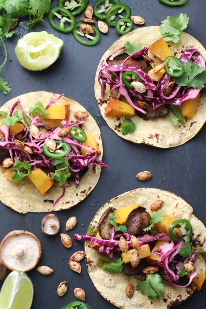 three vegan pumpkin tacos on a dark surface with limes and shiitake mushrooms on the side