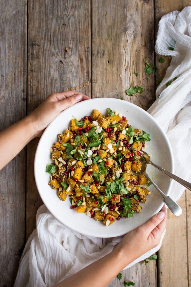 two hands holding a plate of pumpkin salad with pomegranate over a wooden surface 