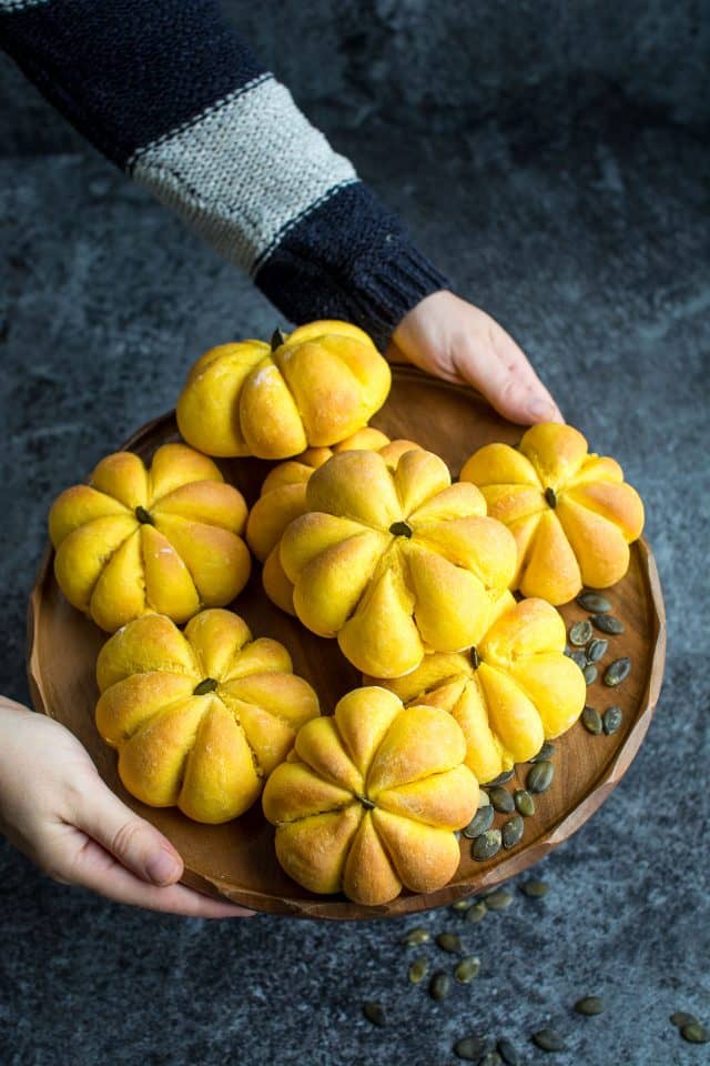 two hands holding a wooden plate with pumpkin rolls over a dark grey surface