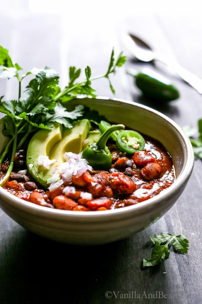 a bowl of pumpkin chili on a wooden counter top with sliced avocado and cilantro on top and a spoon in the background