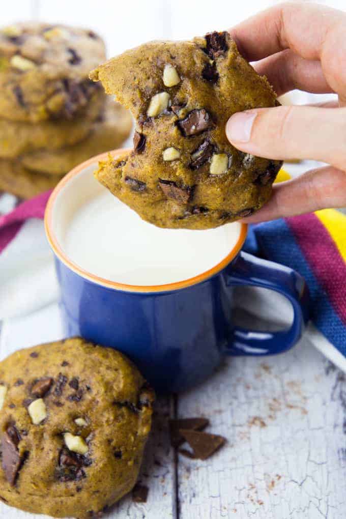 a vegan pumpkin cookie is being hold over a cup of almond milk with more cookies in the background