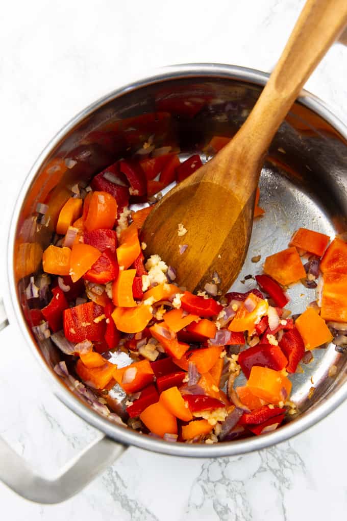 sautéed red onion, garlic, and diced red and orange bell pepper in a pot with a wooden spoon on a marble countertop