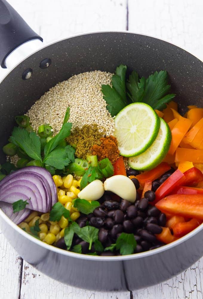 uncooked quinoa, black beans, corn, orange and red bell pepper, and red onion slices in a black pot on a white wooden board 