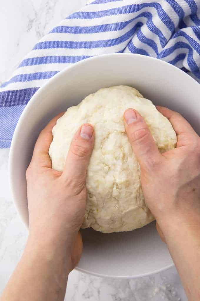 two hands kneading a vegan pizza dough in a white bowl on a marble counter top with a blue dish cloth in the background