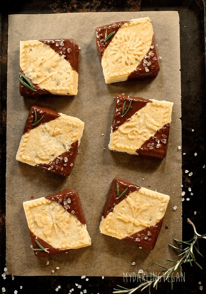 sechs vegane Chocolate-Dipped Shortbread Cookies auf Pergamentpapier auf einer schwarzen Oberfläche mit frischem Rosmarin auf der Seite