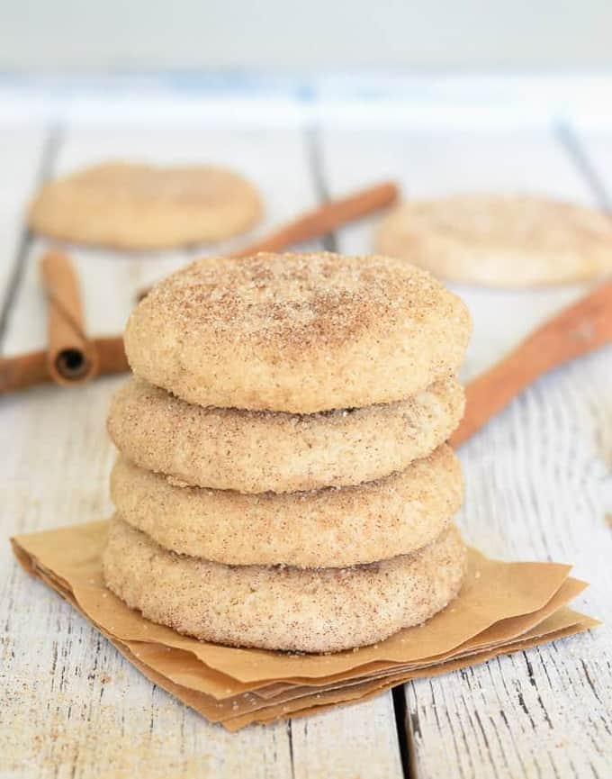 a stack of four vegan snickerdoodle cookies on a white wooden board with cinnamon sticks and more cookies in the background
