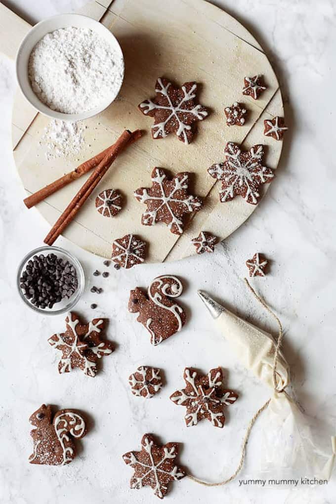 vegan gingerbread cookies in star and squirrel shape with icing on top and two cinnamon sticks, a bowl of powdered sugar, and chocolate chips on the side 