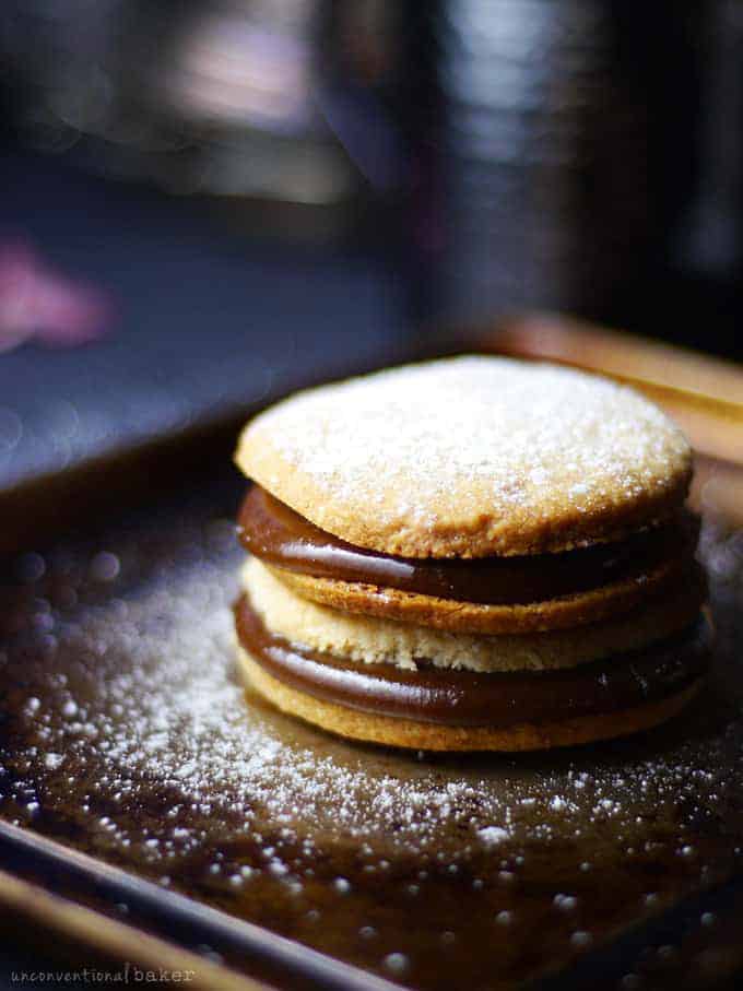 a stack of two vegan alfajores cookies dusted with powdered sugar on a dark plate 