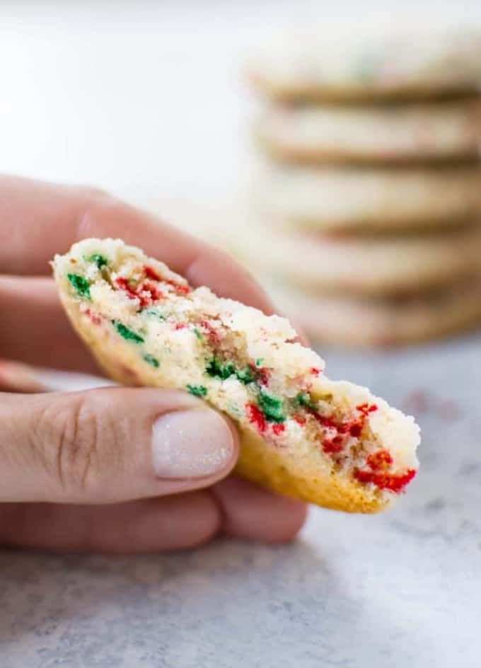 a hand holding a vegan sugar cookie with red and green food coloring with a stack of cookies in the background 