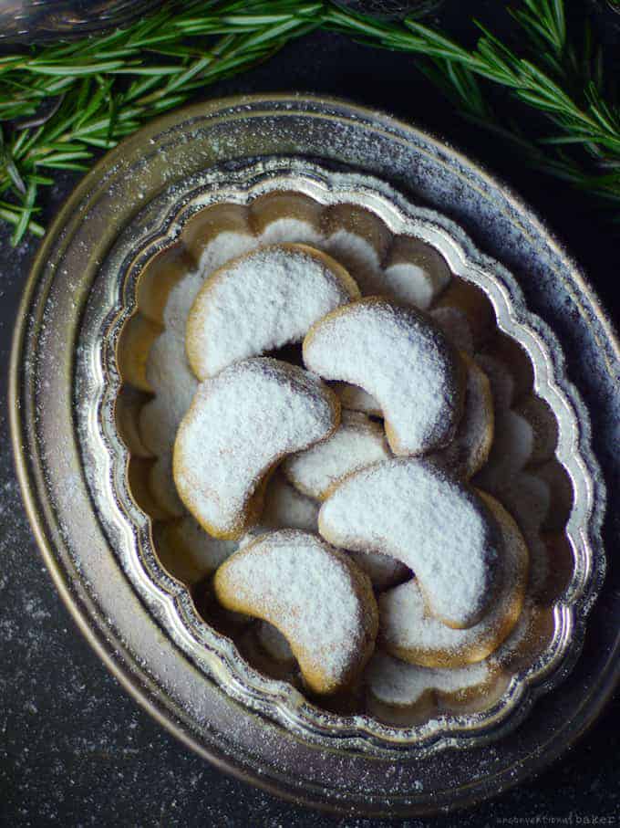 almond crescent cookies med pulverisert sukker på en serveringsplate på en mørk overflate med evergreens i bakgrunnen