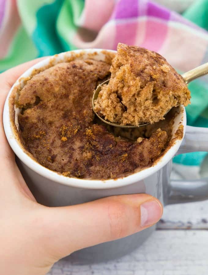 a hand holding a mug with a vegan mug cake on a white wooden board with a green and pink napkin in the background