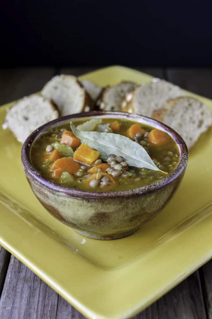 Greek lentil soup in a brown bowl on a yellow plate with slices of bread in the background