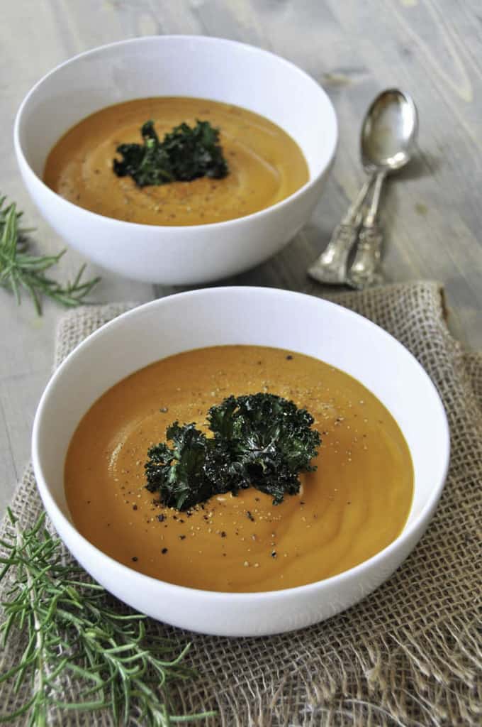 two bowls of vegan carrot soup on a wooden counter top with fresh rosemary and a spoon on the side 