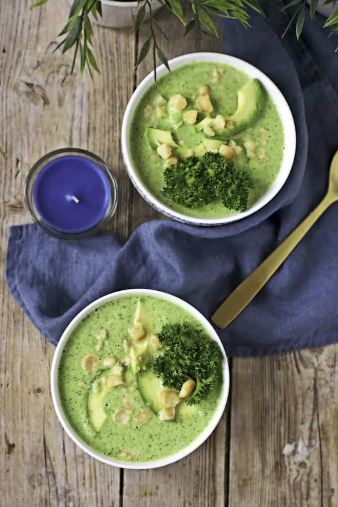 two bowls of vegan broccoli soup on a wooden counter top with a blue dish cloth and a spoon 