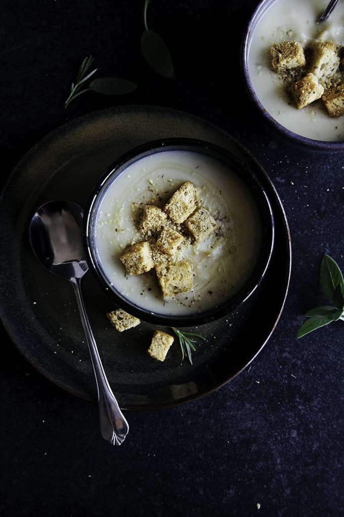 parsnip celeriac soup in two black bowls with crouton on top and a spoon on the side on a dark surface 