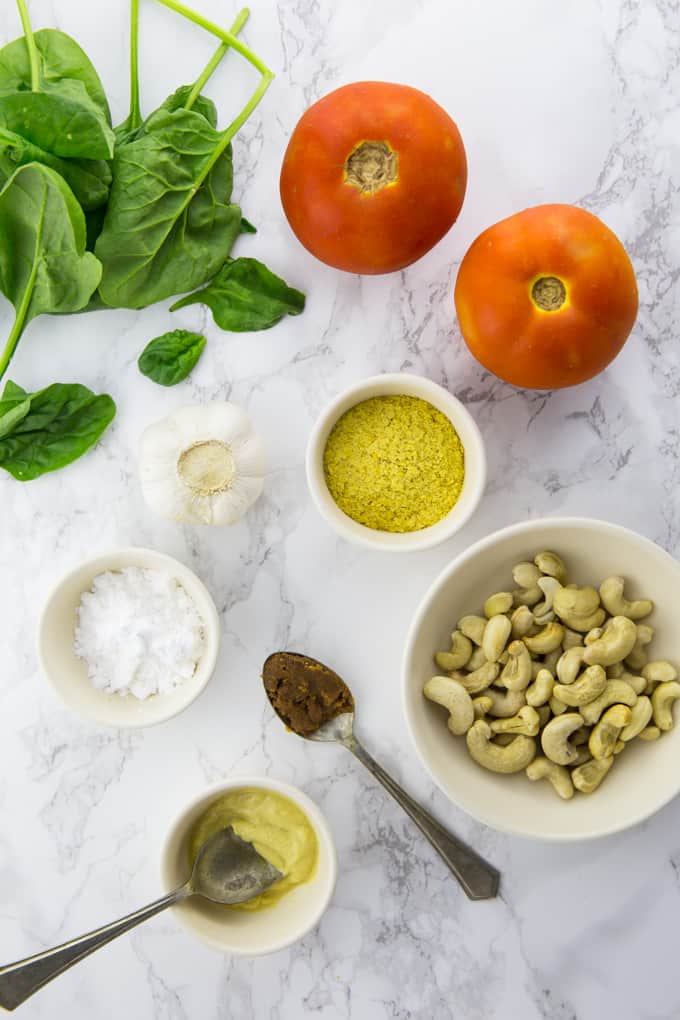 tomatoes, spinach, cashews, nutritional yeast, mustard, garlic, and tapioca starch in small bowls on a marble counter top