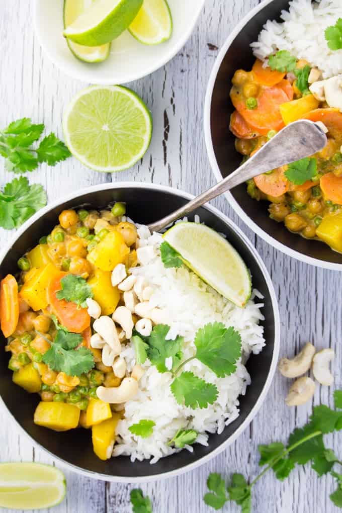 two bowls of vegan chickpea curry on a white wooden surface with cilantro, cashews, and a small bowl of lime wedges on the side 