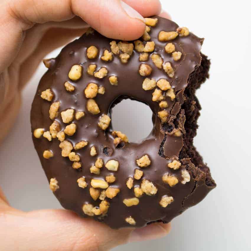 a hand holding a vegan chocolate donut with a missing bite in front of a white wall 
