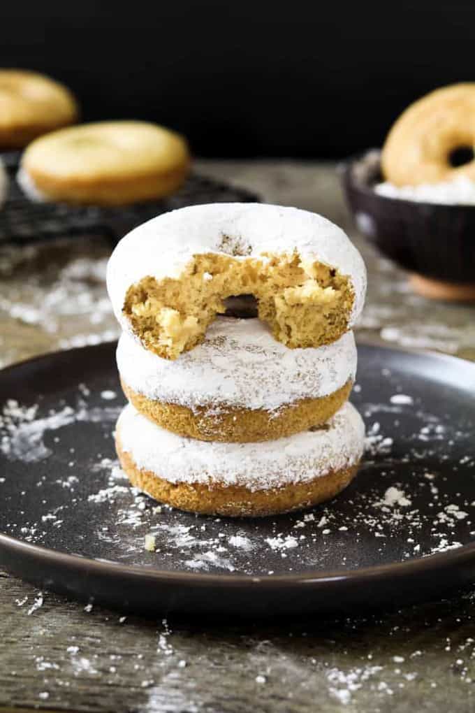 a stack of three vegan powdered donuts on a black plate with more donuts in the background 
