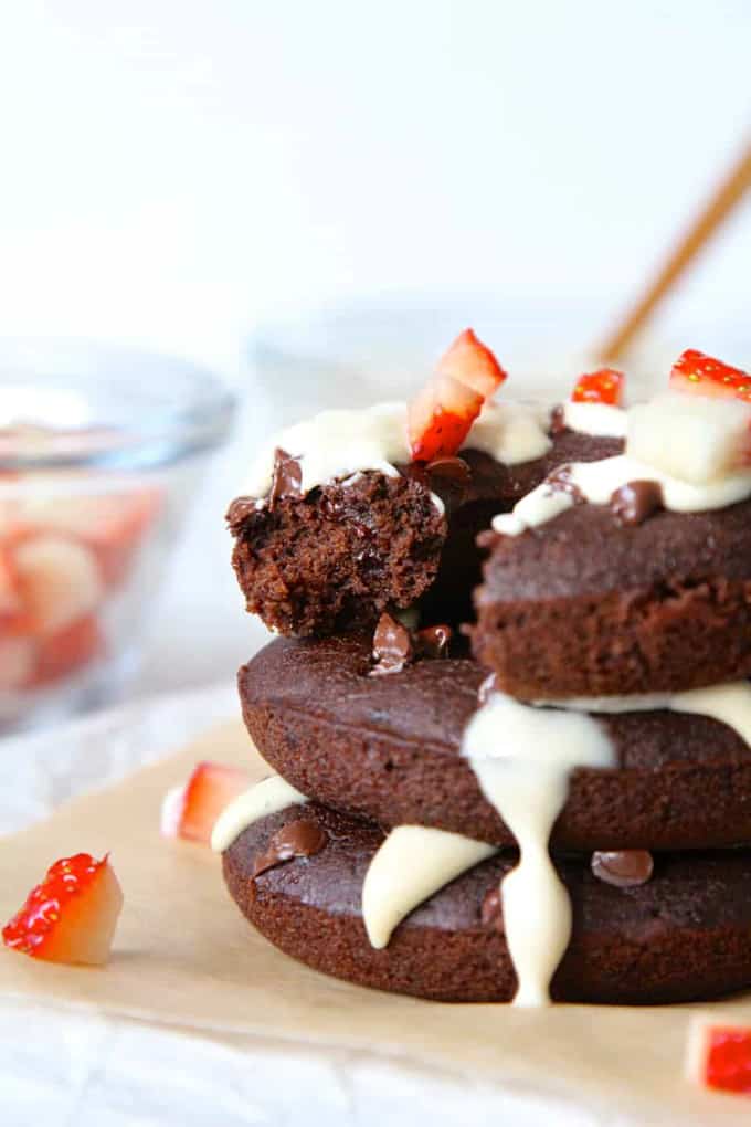 a stack of three vegan chocolate donuts with cashew cream and strawberries on a white counter top with two bowls in the background 