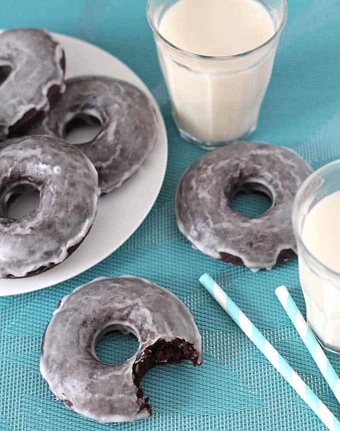 three vegan chocolate donuts on a white plate on a blue dish cloth with two glasses of milk, two donuts, and two blue straws on the side 