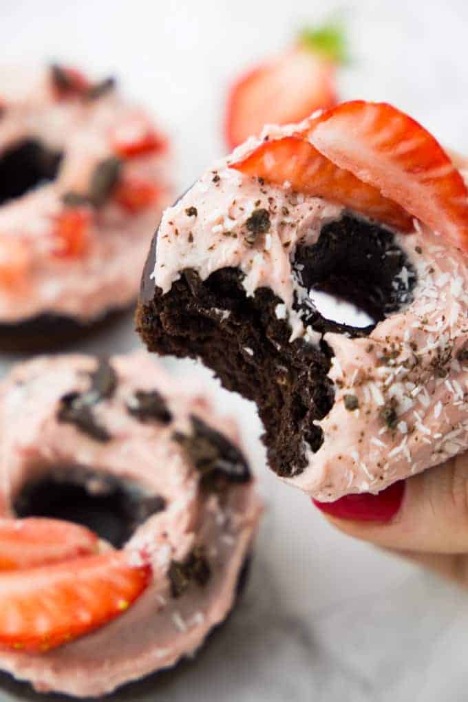 a hand holding a vegan chocolate donut with strawberry frosting with two other donuts and a strawberry in the background 